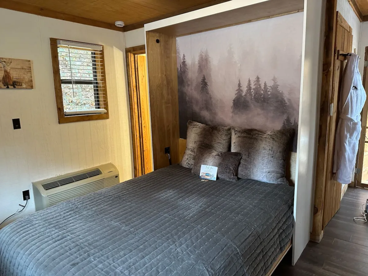 A plush queen-size bed with soft pillows sits against a striking forest mural, with warm wooden accents and artwork on the wall enhancing the cozy, woodland theme. Natural light filters in, and the space feels serene and inviting—ideal for rest and romance.
