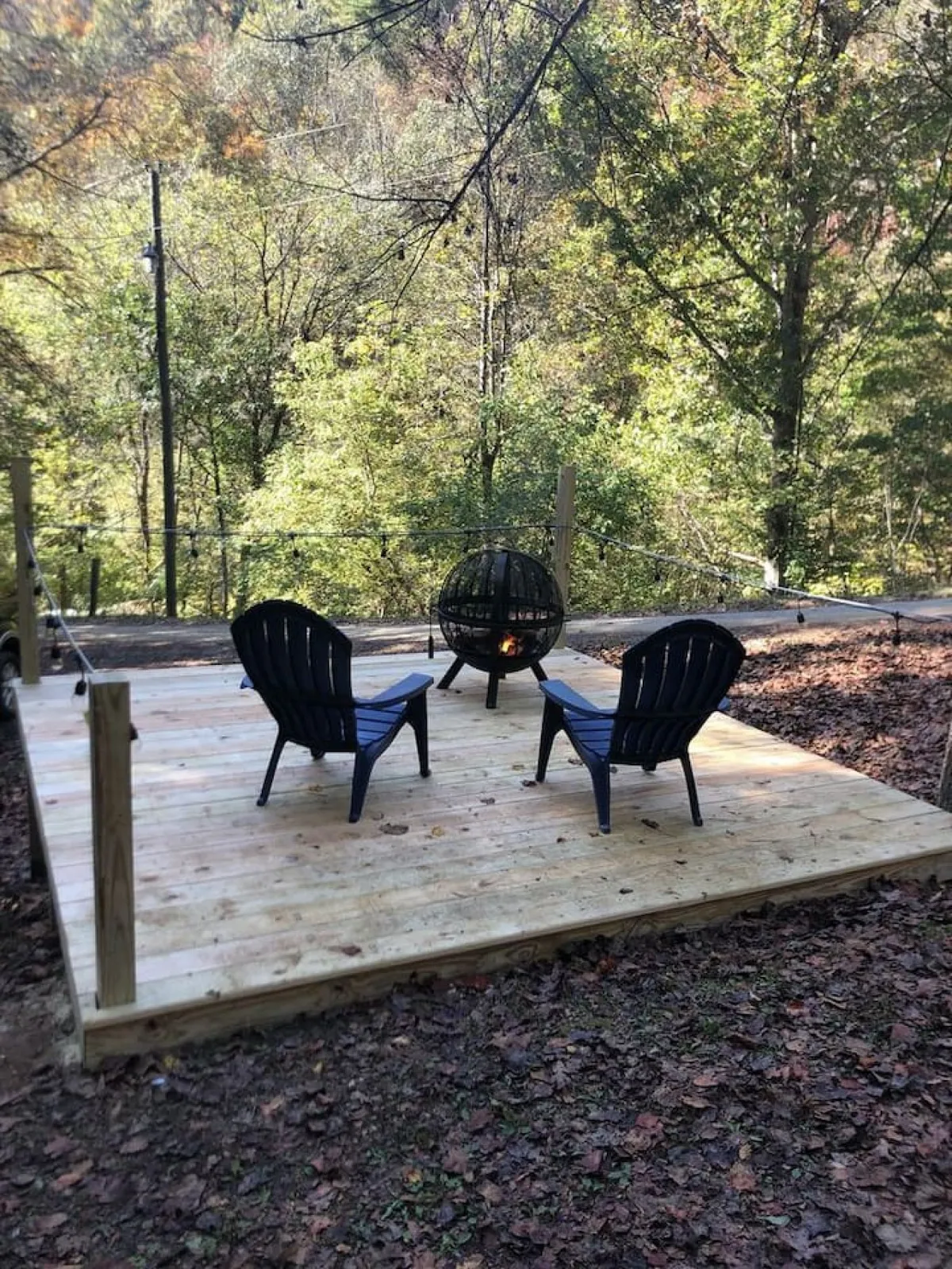 This image features a wooden deck surrounded by trees, with black chairs arranged around a central fire pit. The setting exudes coziness and invites gatherings, with hints of sunset light filtering through the foliage.
