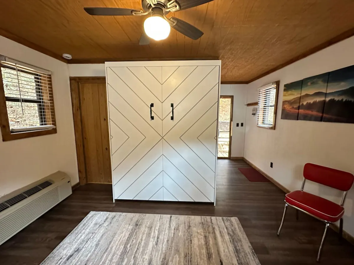 A cozy and efficient interior featuring a white chevron-patterned storage unit, a vibrant red chair, wall art depicting a mountain landscape, and a ceiling fan overhead. Large windows fill the space with natural light, offering glimpses of the outdoors and creating a connection to the natural setting beyond.
