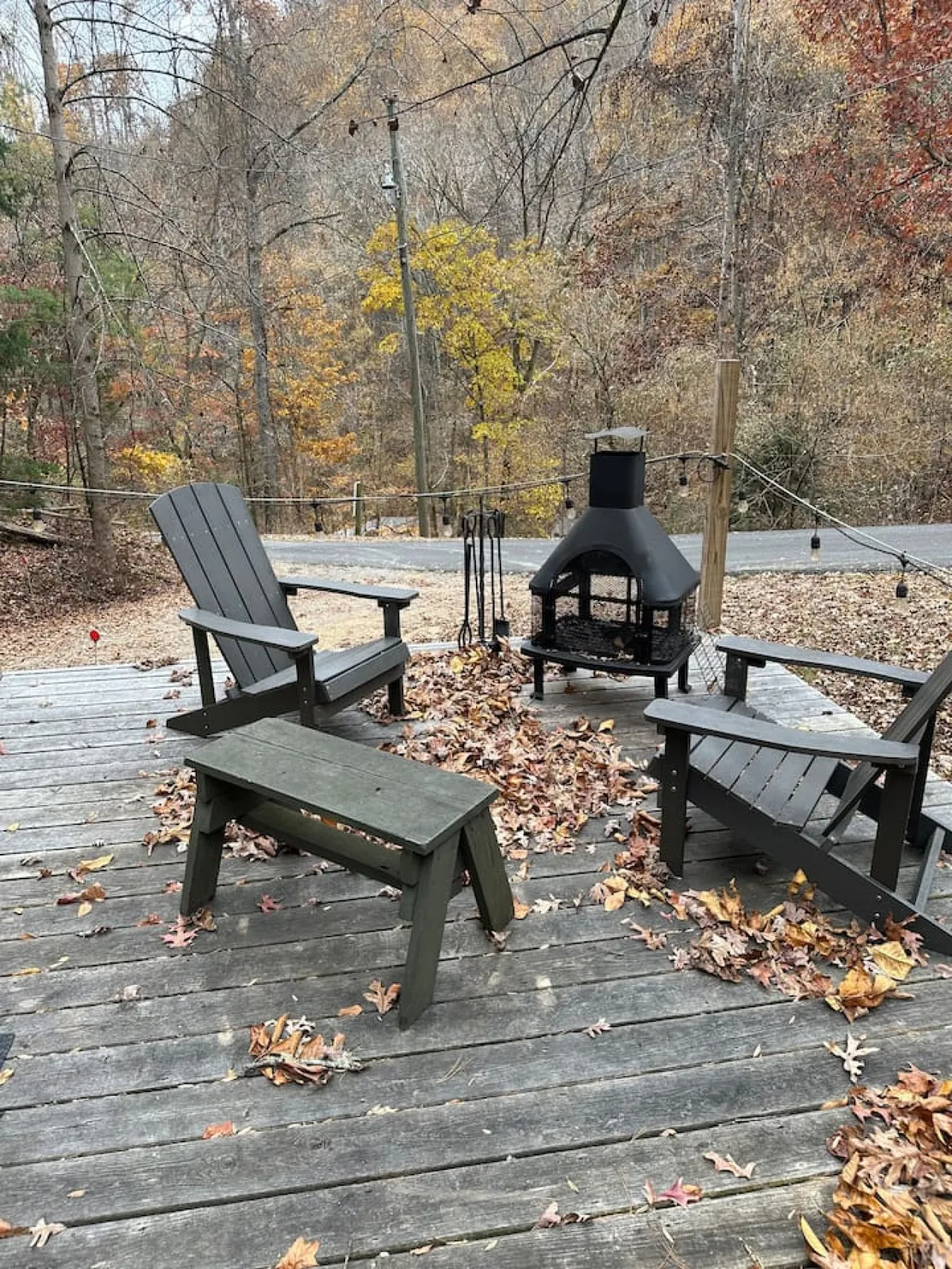 The image displays a welcoming outdoor seating area with classic wooden chairs arranged around a cozy firepit, set amidst fallen leaves and surrounded by tall trees. The serene natural setting emphasizes a sense of peace and invitation to relax outdoors.