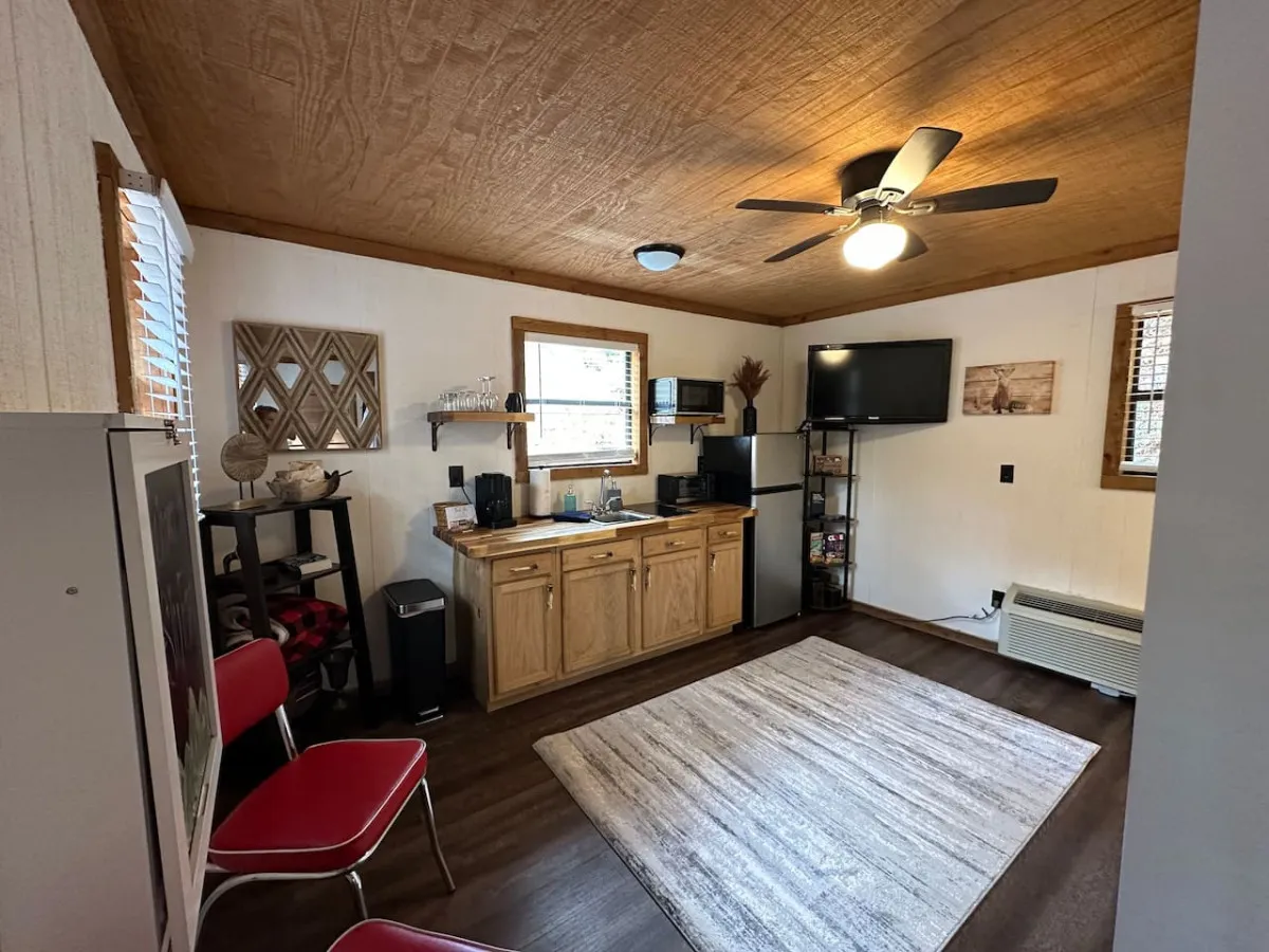 A view of the kitchenette with red chairs, rug, and visible TV, giving a sense of the kitchen’s integration with the living area. The natural light, accessible countertop, and casual seating further highlight the room’s versatility for both dining and relaxing.