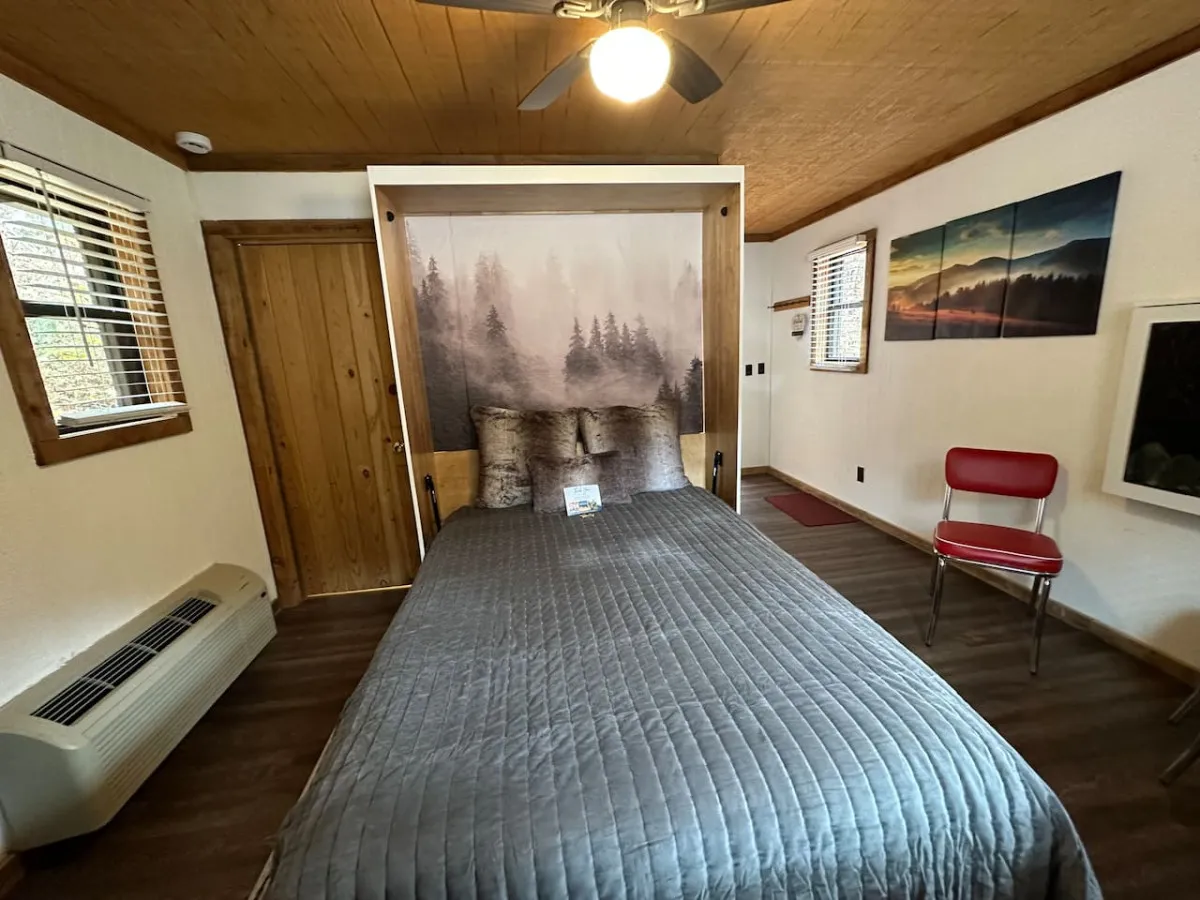 A cozy bed covered with a quilt sits in front of a calming forest mural, accompanied by a comfortable red chair. The room is softly lit, highlighting the tranquil and intimate atmosphere.